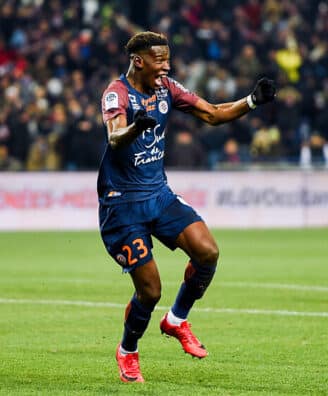 Soccer player in a navy kit (#23) celebrates a goal on a green field as teammates and fans cheer in the background.