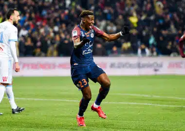Soccer player in a navy kit (#23) celebrates a goal on a green field as teammates and fans cheer in the background.
