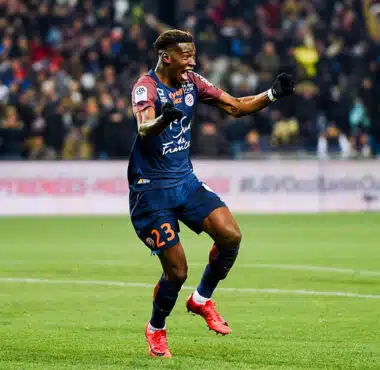Soccer player in a navy kit (#23) celebrates a goal on a green field as teammates and fans cheer in the background.