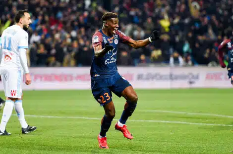 Soccer player in a navy kit (#23) celebrates a goal on a green field as teammates and fans cheer in the background.