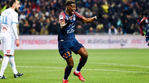 Soccer player in a navy kit (#23) celebrates a goal on a green field as teammates and fans cheer in the background.