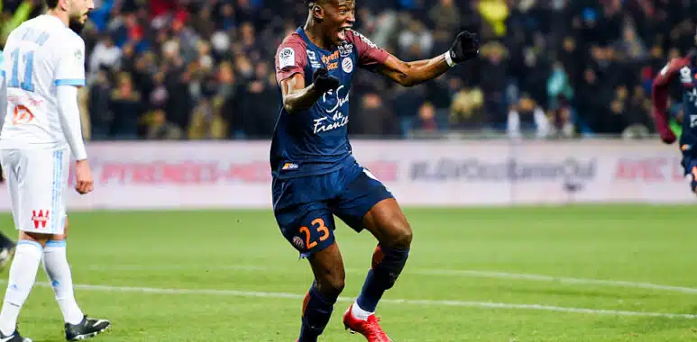 Nordi Mukiele | AllezPaillade.com Soccer player in a navy kit (#23) celebrates a goal on a green field as teammates and fans cheer in the background.