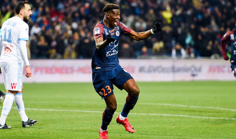 Soccer player in a navy kit (#23) celebrates a goal on a green field as teammates and fans cheer in the background.