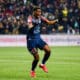 Soccer player in a navy kit (#23) celebrates a goal on a green field as teammates and fans cheer in the background.