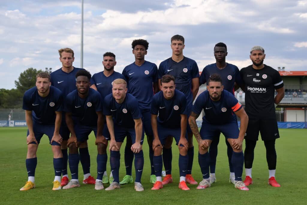 Eleven soccer players pose for a team photo on a green field, wearing matching navy kits with a crest; a goalkeeper in black stands on the right.