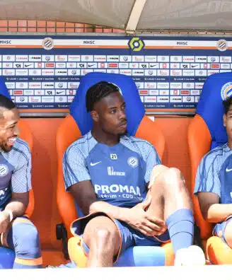 Three soccer players sit on a dugout bench in blue kits with sponsor logos, chatting during a break against a backdrop of sponsor boards.