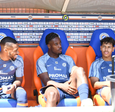Three soccer players sit on a dugout bench in blue kits with sponsor logos, chatting during a break against a backdrop of sponsor boards.