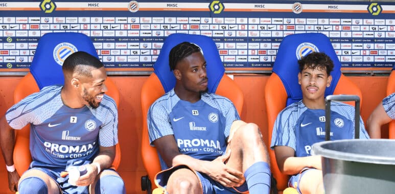 Three soccer players sit on a dugout bench in blue kits with sponsor logos, chatting during a break against a backdrop of sponsor boards.