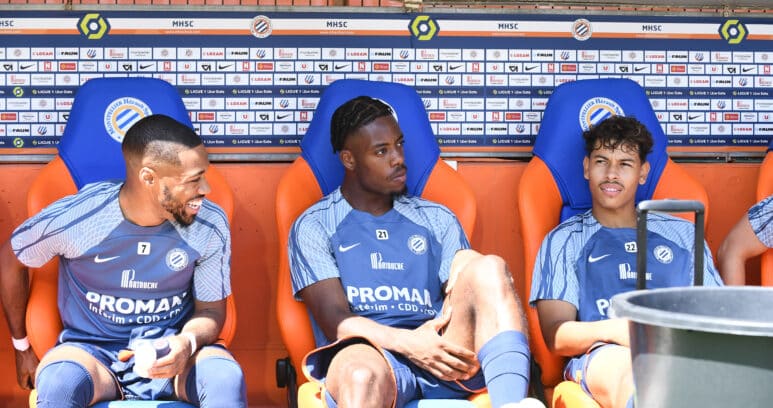 Three soccer players sit on a dugout bench in blue kits with sponsor logos, chatting during a break against a backdrop of sponsor boards.