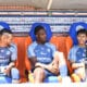 Three soccer players sit on a dugout bench in blue kits with sponsor logos, chatting during a break against a backdrop of sponsor boards.