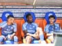 Three soccer players sit on a dugout bench in blue kits with sponsor logos, chatting during a break against a backdrop of sponsor boards.