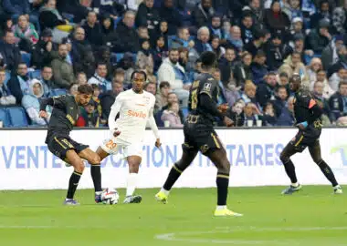 Soccer players chase the ball on a green pitch with a crowded stands in the background.