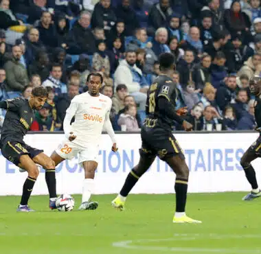 Soccer players chase the ball on a green pitch with a crowded stands in the background.