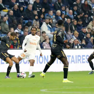 Soccer players chase the ball on a green pitch with a crowded stands in the background.