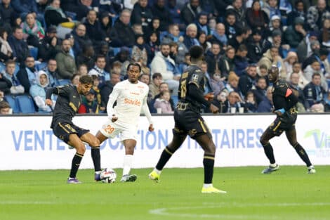 Soccer players chase the ball on a green pitch with a crowded stands in the background.