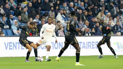 Soccer players chase the ball on a green pitch with a crowded stands in the background.