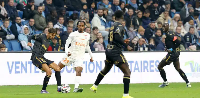 Soccer players chase the ball on a green pitch with a crowded stands in the background.