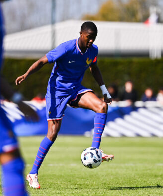 Soccer player in blue jersey controlling a ball on a grassy field with teammates and spectators in the background