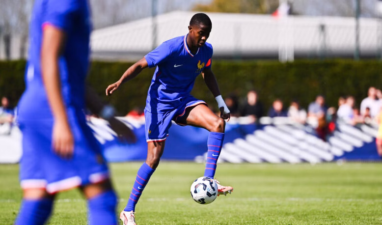 Soccer player in blue jersey controlling a ball on a grassy field with teammates and spectators in the background