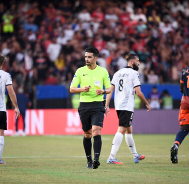 Soccer match scene with a neon-clad referee at center and players around him on a green field with a crowded stadium in the background.