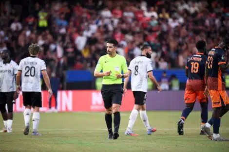 Soccer match scene with a neon-clad referee at center and players around him on a green field with a crowded stadium in the background.