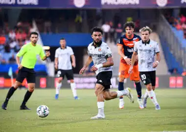 Soccer match scene: a player in white jersey dribbles the ball on the field as teammates and opponents chase behind him.