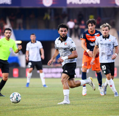 Soccer match scene: a player in white jersey dribbles the ball on the field as teammates and opponents chase behind him.