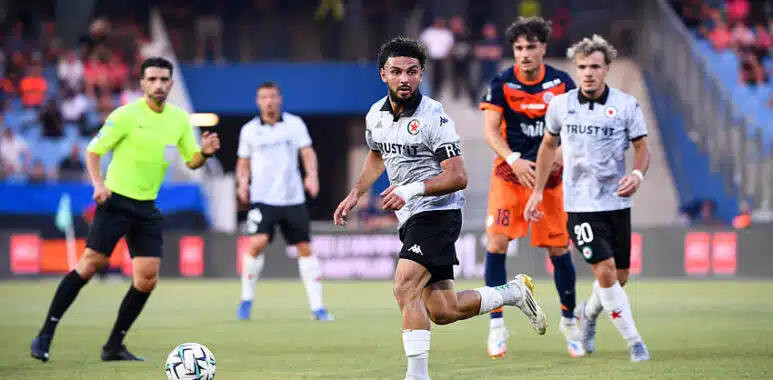 Ryad Hachem | AllezPaillade.com Soccer match scene: a player in white jersey dribbles the ball on the field as teammates and opponents chase behind him.