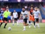 Soccer match scene: a player in white jersey dribbles the ball on the field as teammates and opponents chase behind him.
