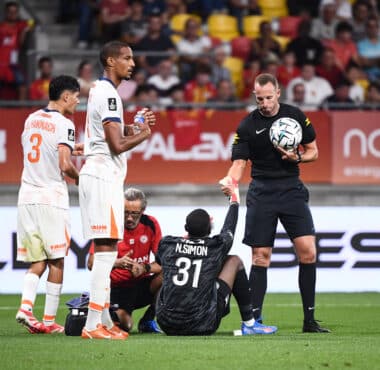 Soccer scene: a goalkeeper in black kneels on the ground holding a ball while two players in white uniforms stand nearby on a green pitch, with a crowded stadium in the background.