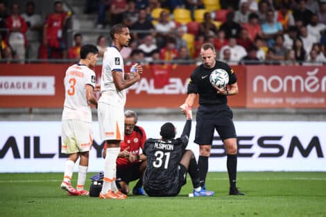 Soccer scene: a goalkeeper in black kneels on the ground holding a ball while two players in white uniforms stand nearby on a green pitch, with a crowded stadium in the background.