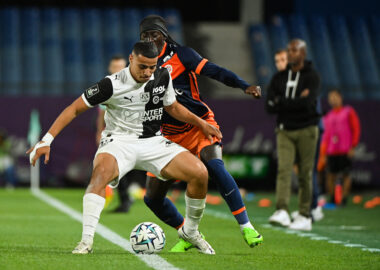 Two soccer players clash for the ball on a green field, one in a white kit and the other in navy with orange accents, both focused on the ball