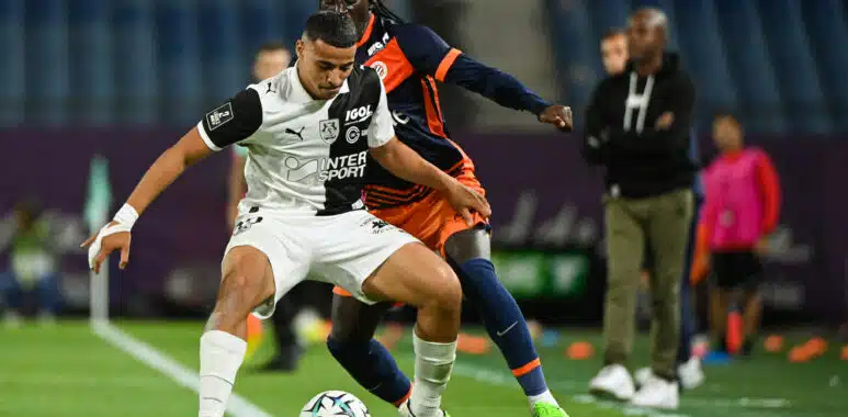 Two soccer players clash for the ball on a green field, one in a white kit and the other in navy with orange accents, both focused on the ball