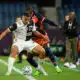 Two soccer players clash for the ball on a green field, one in a white kit and the other in navy with orange accents, both focused on the ball