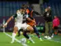 Two soccer players clash for the ball on a green field, one in a white kit and the other in navy with orange accents, both focused on the ball