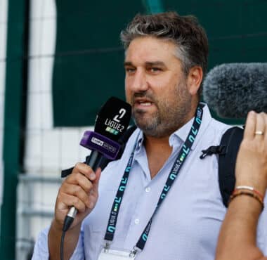 Man being interviewed by reporters, speaking into a microphone labeled Ligue 2, with a second microphone held nearby.