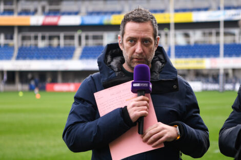 Male sports reporter in a navy jacket holds a purple microphone and pink clipboard on a stadium field.