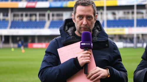 Male sports reporter in a navy jacket holds a purple microphone and pink clipboard on a stadium field.