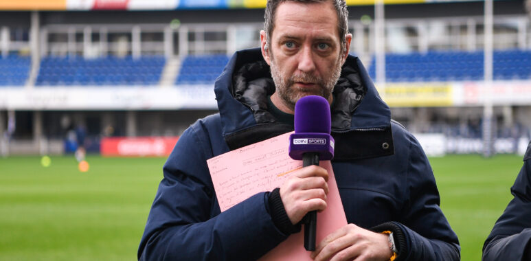 Male sports reporter in a navy jacket holds a purple microphone and pink clipboard on a stadium field.