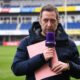 Male sports reporter in a navy jacket holds a purple microphone and pink clipboard on a stadium field.