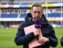Male sports reporter in a navy jacket holds a purple microphone and pink clipboard on a stadium field.