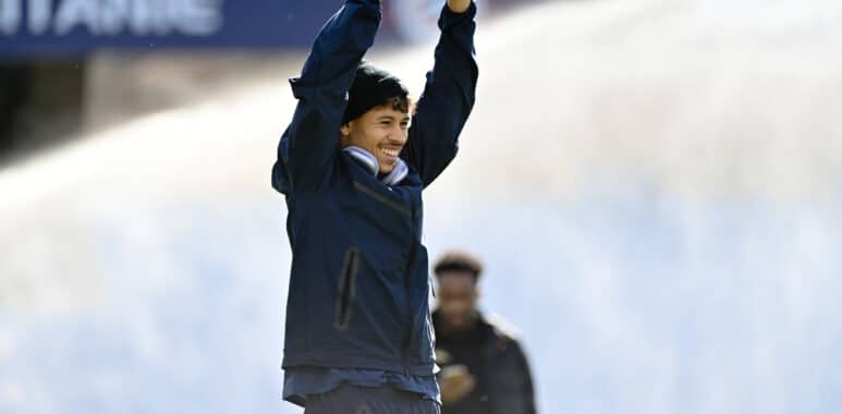Man in a navy jacket raises arms in celebration on a sports field, smiling at the camera.