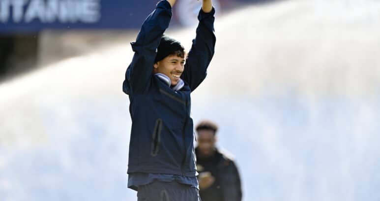 Man in a navy jacket raises arms in celebration on a sports field, smiling at the camera.