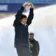 Man in a navy jacket raises arms in celebration on a sports field, smiling at the camera.