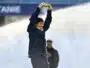 Man in a navy jacket raises arms in celebration on a sports field, smiling at the camera.