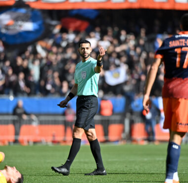 Soccer referee in a mint-green shirt raises a hand as players in orange navy jerseys stand nearby; a fallen yellow-clad player lies on the field.