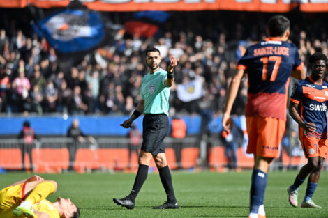 Soccer referee in a mint-green shirt raises a hand as players in orange navy jerseys stand nearby; a fallen yellow-clad player lies on the field.