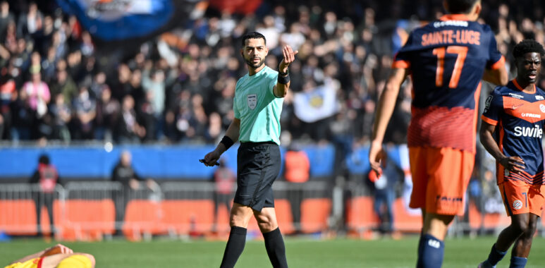 Soccer referee in a mint-green shirt raises a hand as players in orange navy jerseys stand nearby; a fallen yellow-clad player lies on the field.