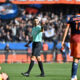 Soccer referee in a mint-green shirt raises a hand as players in orange navy jerseys stand nearby; a fallen yellow-clad player lies on the field.