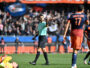 Soccer referee in a mint-green shirt raises a hand as players in orange navy jerseys stand nearby; a fallen yellow-clad player lies on the field.